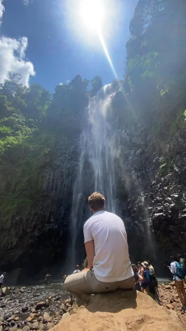 A visitor sitting on a rock at the base of Materuni Falls with sunlight over the cliff